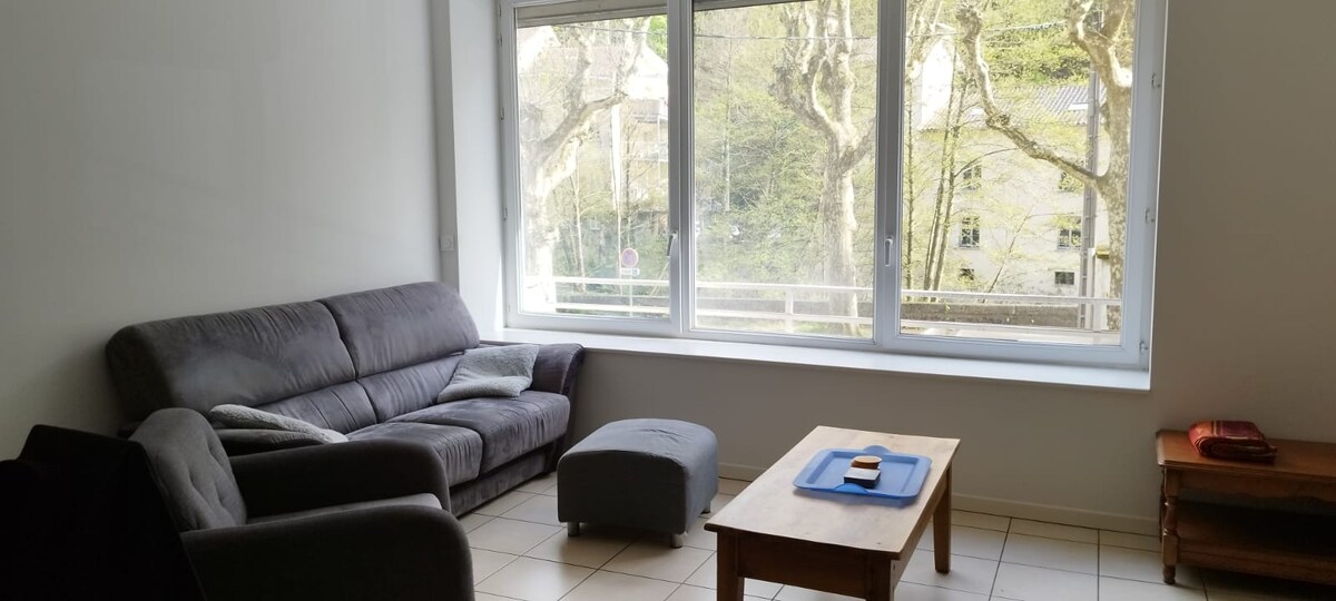 A spacious living area is featured, highlighted by large windows that allow natural light to fill the room. A comfortable gray sofa is positioned alongside an ottoman and a wooden coffee table. A simple cabinet is placed against the wall, contributing to the uncluttered atmosphere.