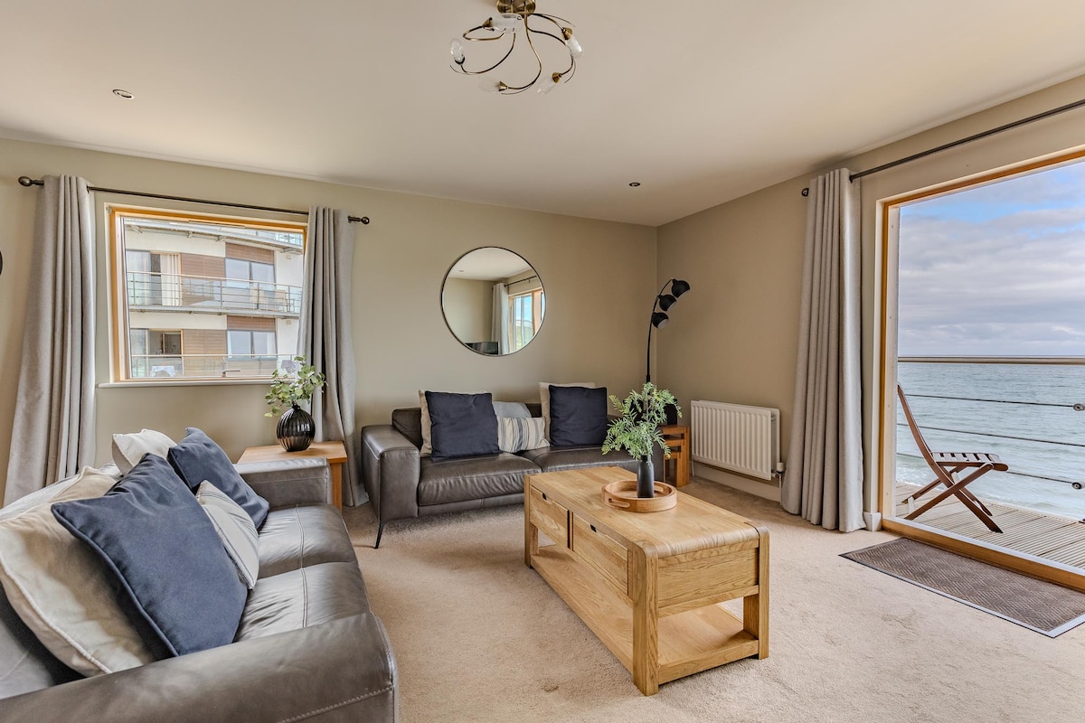 A light-filled living area features two windows offering views of the sea. A round mirror reflects natural light, while a grey sofa with decorative pillows and a wooden coffee table create a relaxed setting. Soft curtains frame the windows, complementing the neutral wall tones.