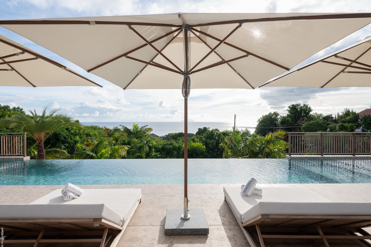 An infinity pool is framed by lush greenery and ocean views. Two loungers are positioned under shade umbrellas, with rolled towels neatly placed on each. The serene landscape creates a calming atmosphere, inviting relaxation and enjoyment of the surroundings.