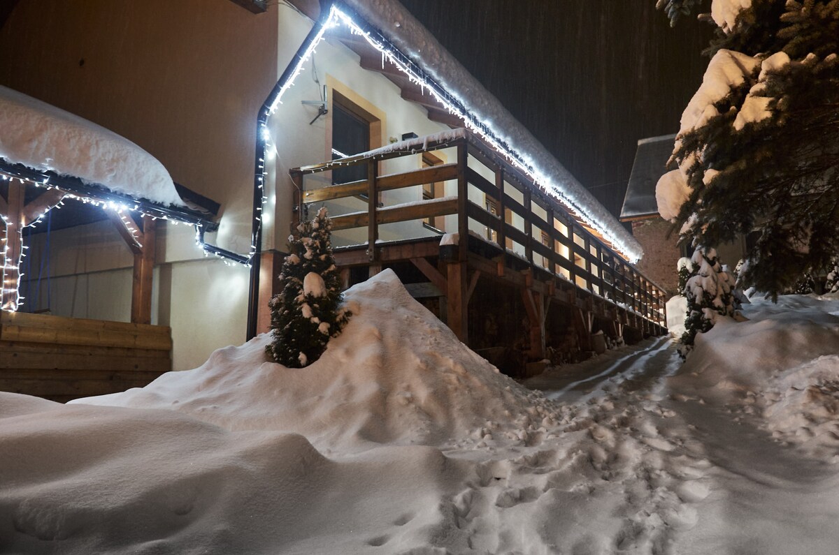 A winter scene shows a charming building adorned with soft white lights, set against a backdrop of falling snow. A wooden balcony stretches across the front, surrounded by freshly fallen snow, with footsteps leading towards the entrance.