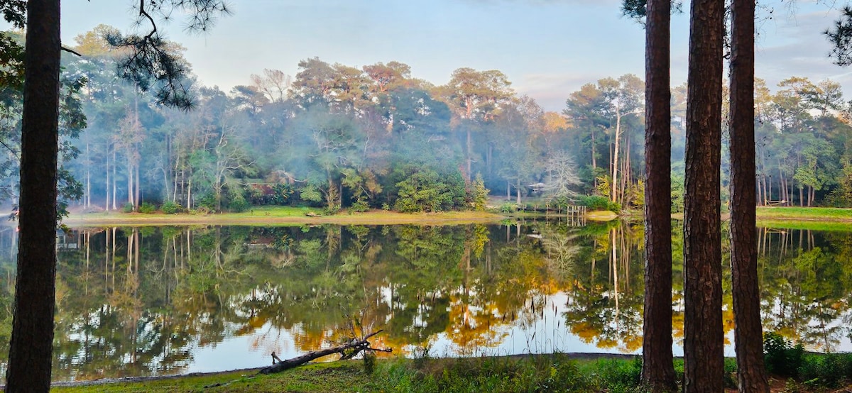 A serene lake is surrounded by tall trees, with mist softly hovering over the water's surface. The reflections of the trees and foliage create a peaceful scene, while a fallen log rests at the water's edge, adding to the natural tranquility.