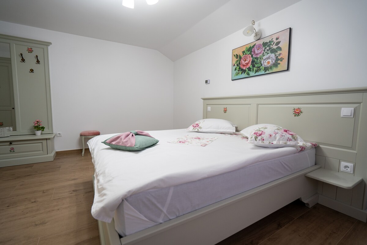 A spacious bedroom features a large bed with floral-patterned pillows and a light duvet. A decorative painting of flowers hangs above the headboard. A wardrobe is visible, and a small stool accompanies the room’s neutral walls and wooden flooring.