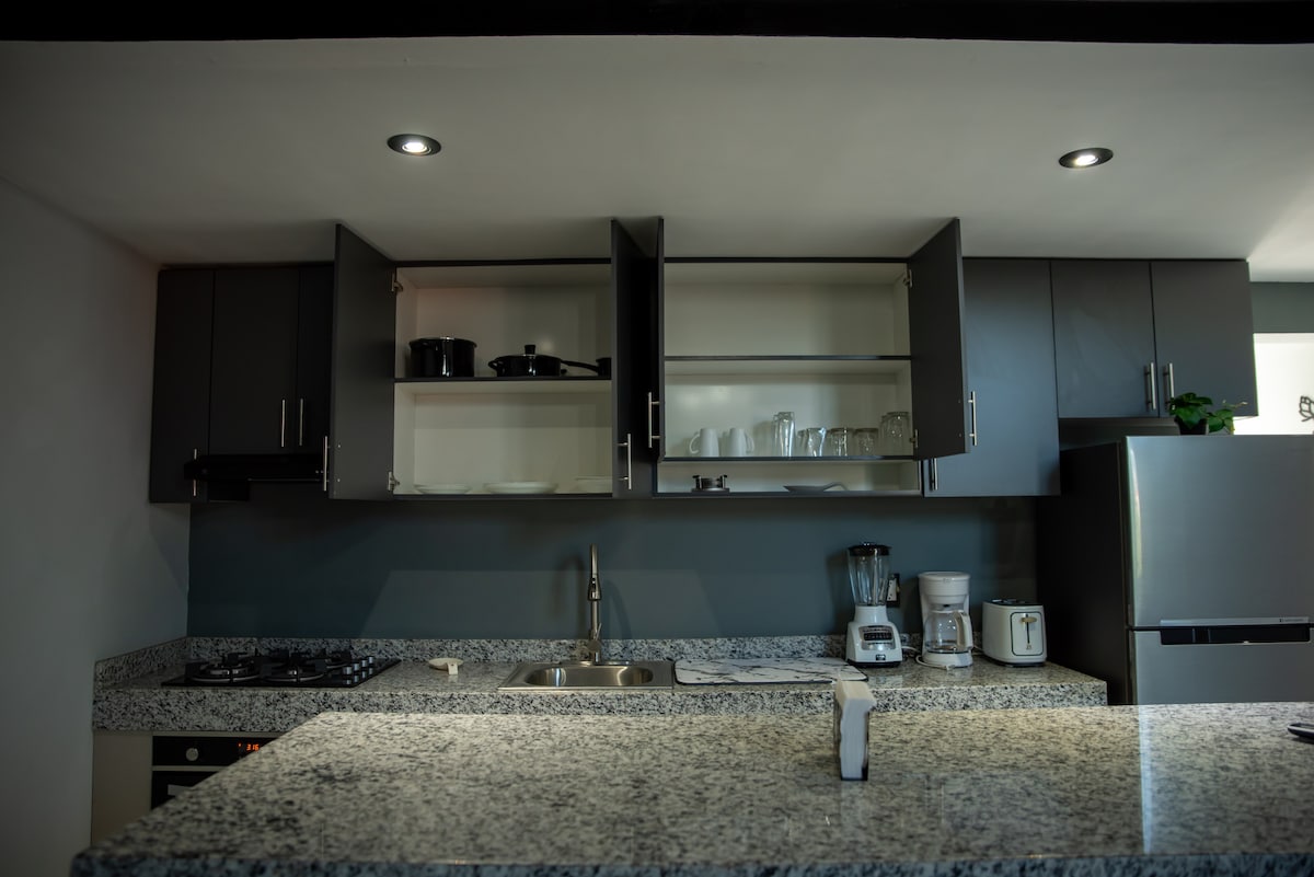 A modern kitchen is presented with sleek cabinets and a granite countertop. Open shelving reveals dishes and glassware, while appliances such as a blender and coffee maker are neatly arranged. Subtle lighting from above enhances the clean, organized look of the space.