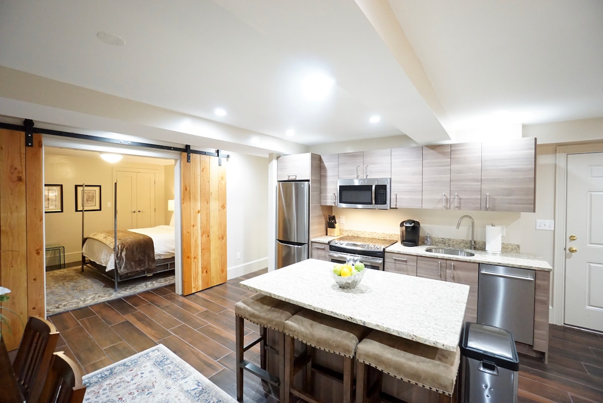 An open kitchen showcases modern stainless steel appliances and a large granite island with three bar stools. Farm doors lead to the bedroom in the background, providing a seamless transition between spaces. Natural light is present throughout the area, enhancing the inviting layout.