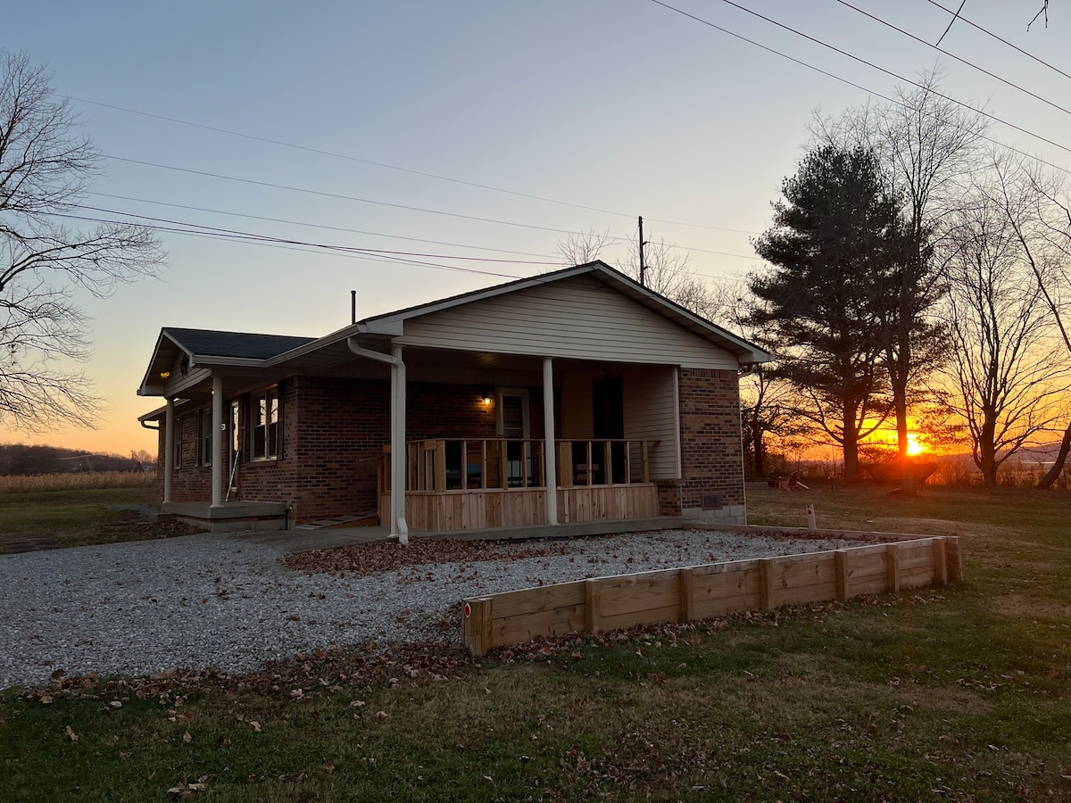 A cozy house is viewed from a distance, showcasing a brick exterior and a welcoming porch. The setting sun creates a serene backdrop, casting warm colors over the landscape. Surrounding greenery complements the natural ambiance of the area.