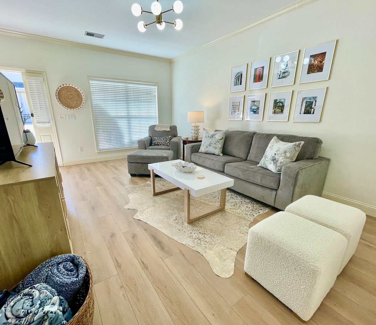 A comfortable living area features a gray sectional sofa and a matching armchair, arranged around a white coffee table. The light-colored rug adds warmth to the space. Decorative artwork adorns the walls, and natural light enters through the window, creating a bright and inviting environment.