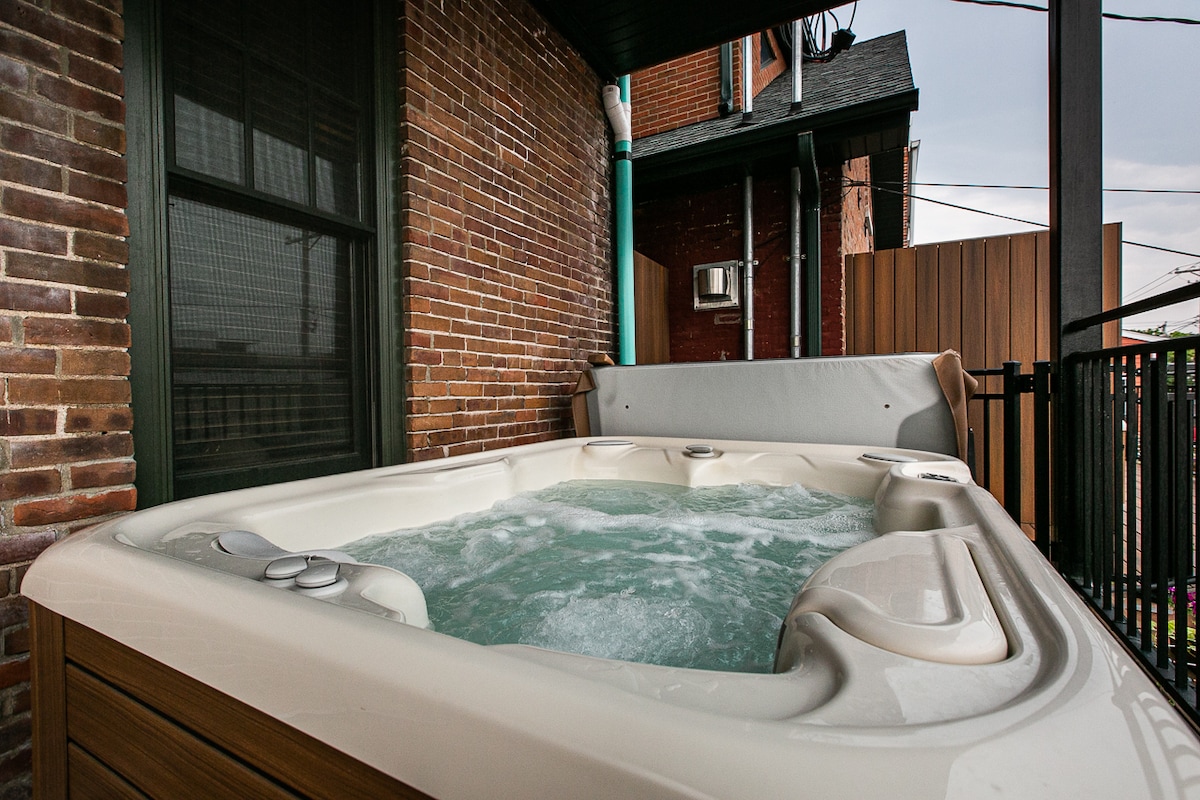 A private hot tub is showcased on the deck, featuring a sleek design with bubbling water and a protective cover. The hot tub is surrounded by a wooden border, complemented by exposed brick walls in the backdrop, enhancing the outdoor space's privacy.
