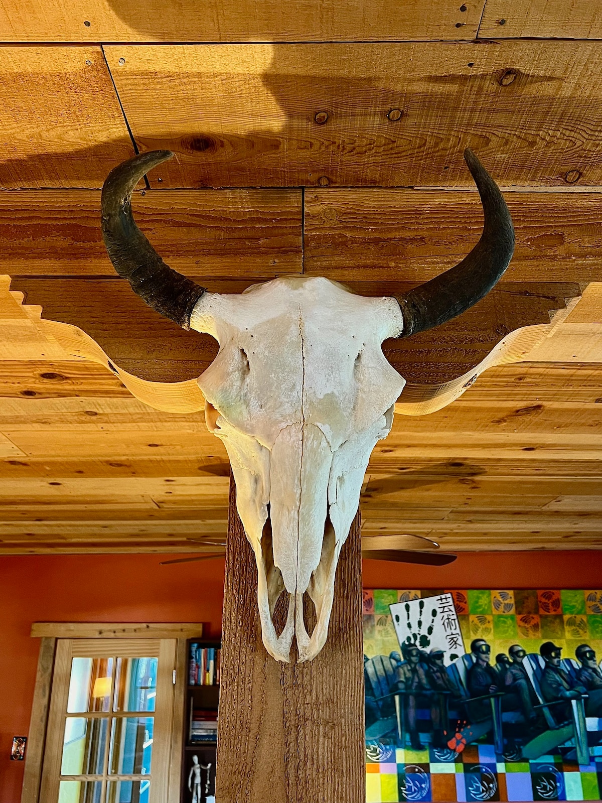 A decorative longhorn skull is mounted on a wooden beam, showcasing its natural color and distinct features. The backdrop includes warm orange walls and an inviting wooden ceiling, contributing to the rustic charm of the space.