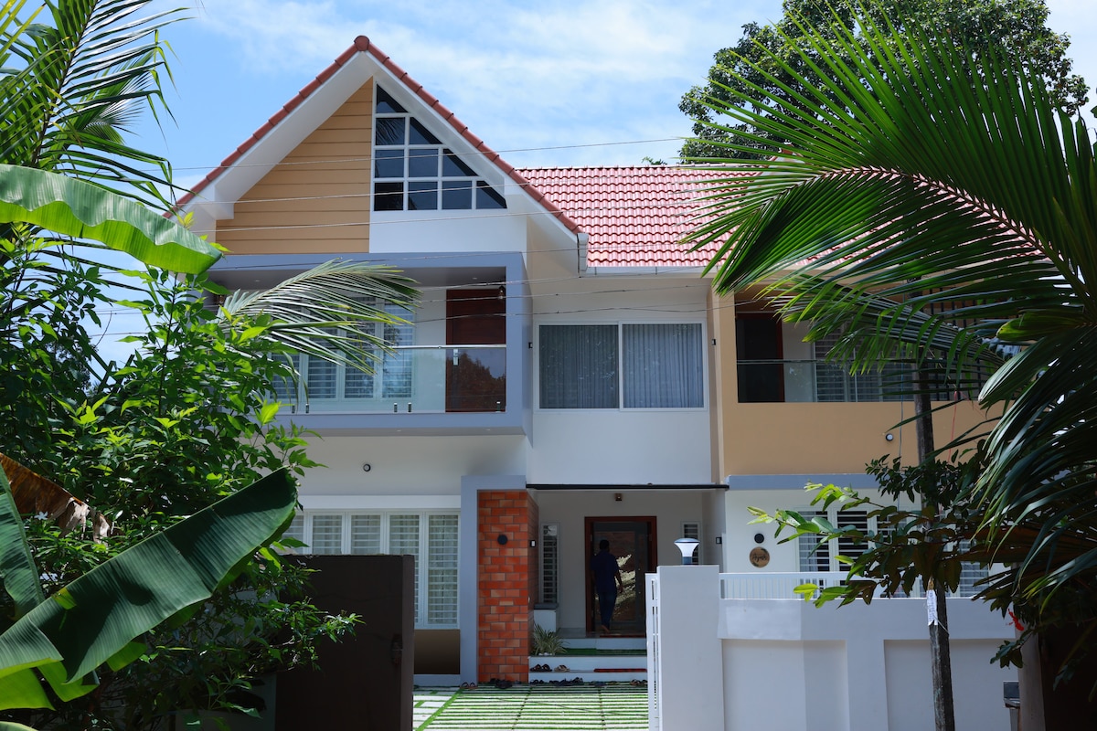 The exterior of a modern villa is viewed, showcasing a blend of white and orange walls with a red-tiled roof. Lush greenery surrounds the entrance, and large windows allow natural light to illuminate the interior. A pathway leads to the main door.