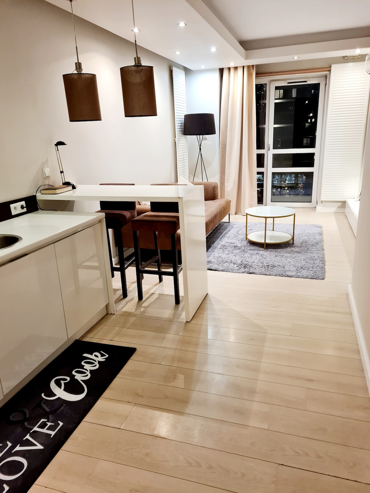 A modern kitchenette features sleek white cabinetry and a small dining area with two dark brown stools. A cozy living space includes a light brown sofa, a round coffee table, and a plush rug, with large windows allowing natural light to fill the room.
