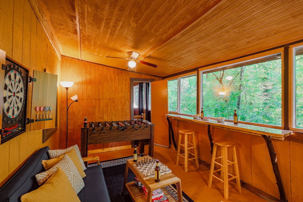 A vibrant entertainment space features warm orange walls, a cozy sofa, and a dartboard. A Foosball table occupies the center, complemented by bar stools at a wooden counter with large windows offering views of the surrounding greenery.