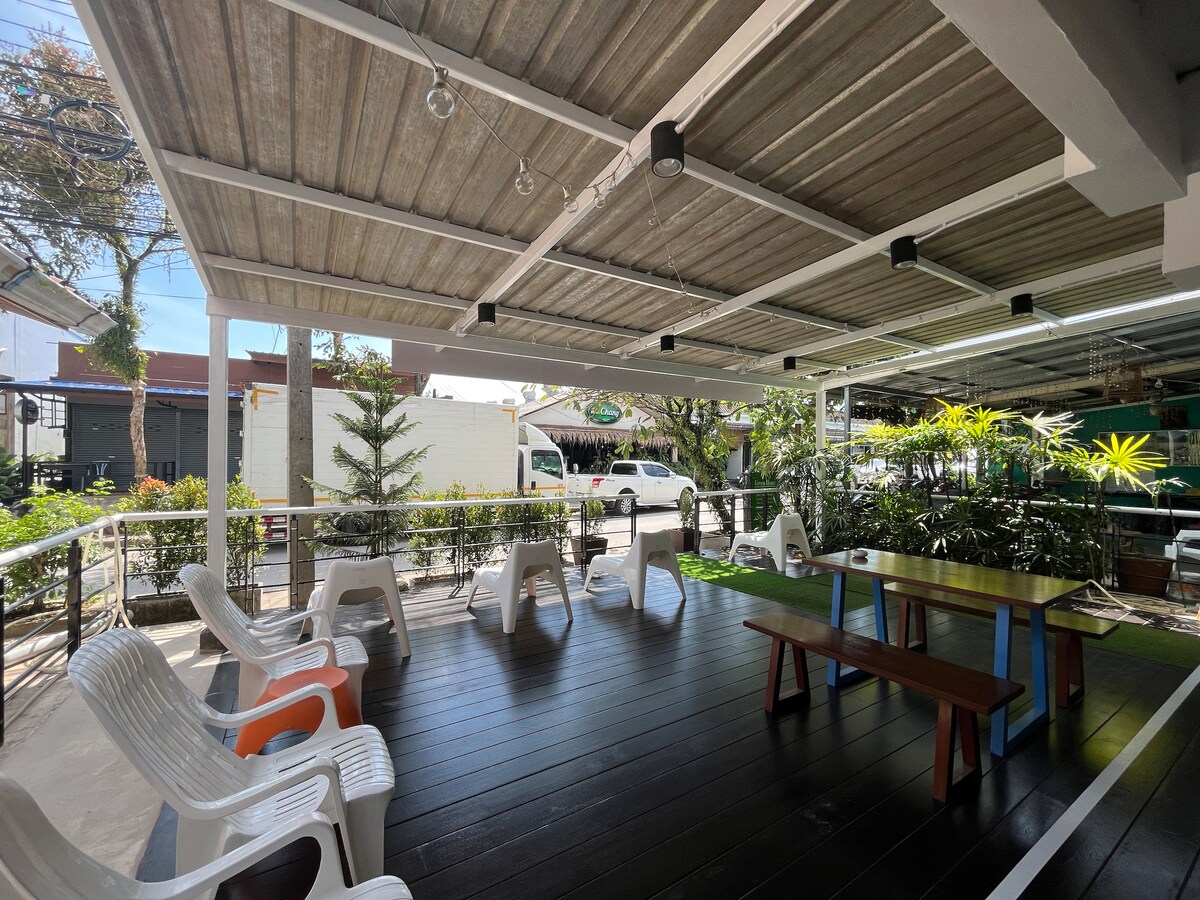 An outdoor seating area features multiple white plastic chairs arranged around a wooden table. The space is shaded by a covered roof and surrounded by greenery, creating a comfortable environment for relaxation. Soft lighting fixtures hang overhead, enhancing the ambiance.