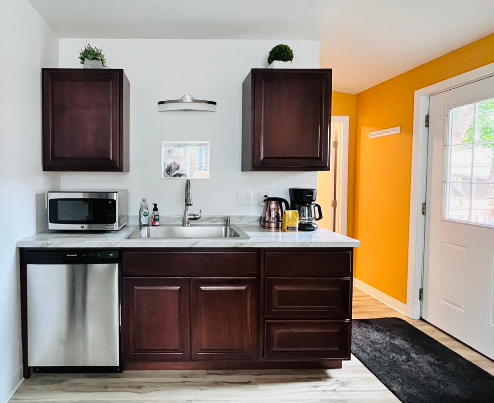 The kitchen area features dark wooden cabinets above a white countertop. A stainless steel sink is centered between a microwave and a coffee maker. A doorway to the right leads to another room, while a bright orange wall creates a warm contrast to the space.