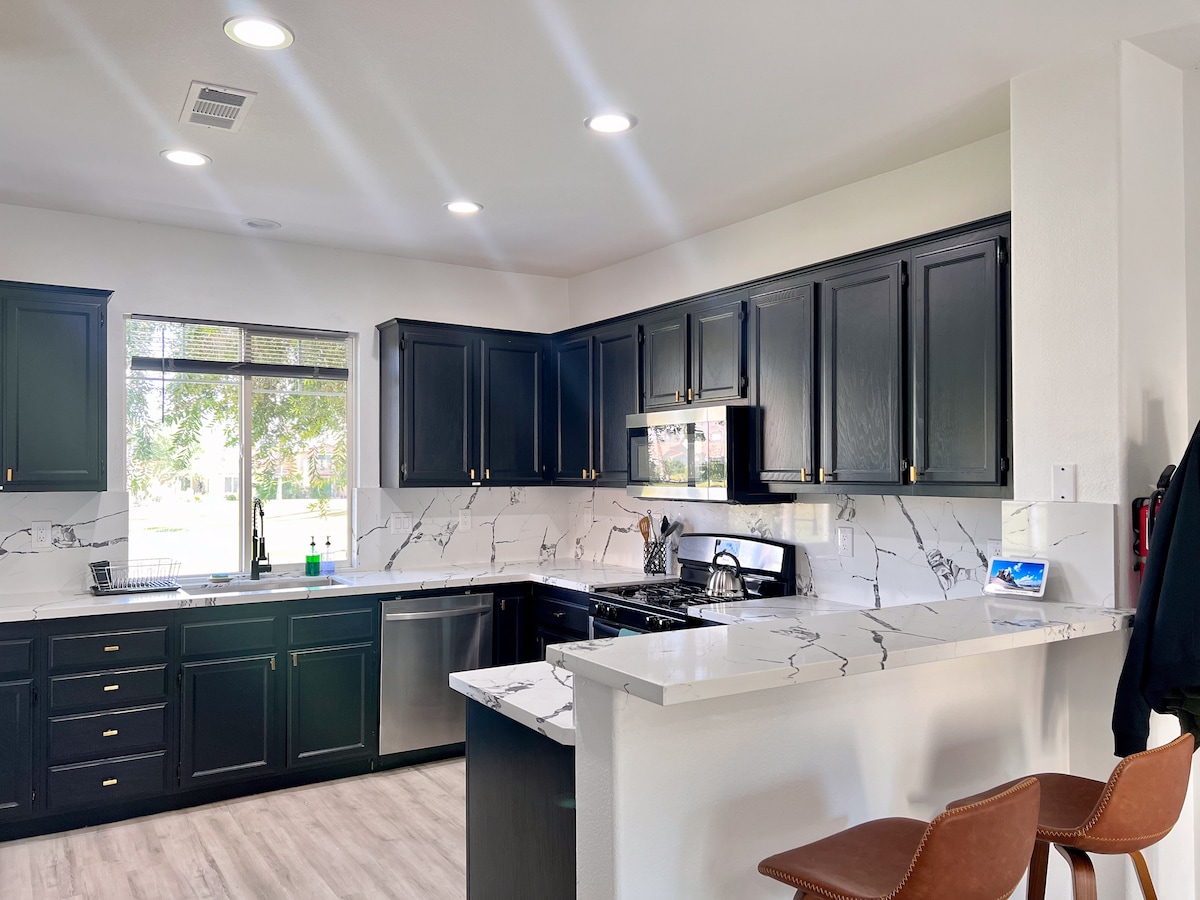 The modern kitchen features sleek black cabinetry, complemented by a white marble countertop. Stainless steel appliances are included, and a window allows natural light to fill the space, highlighting the contemporary design. Two stylish bar stools are positioned at the countertop for seating.