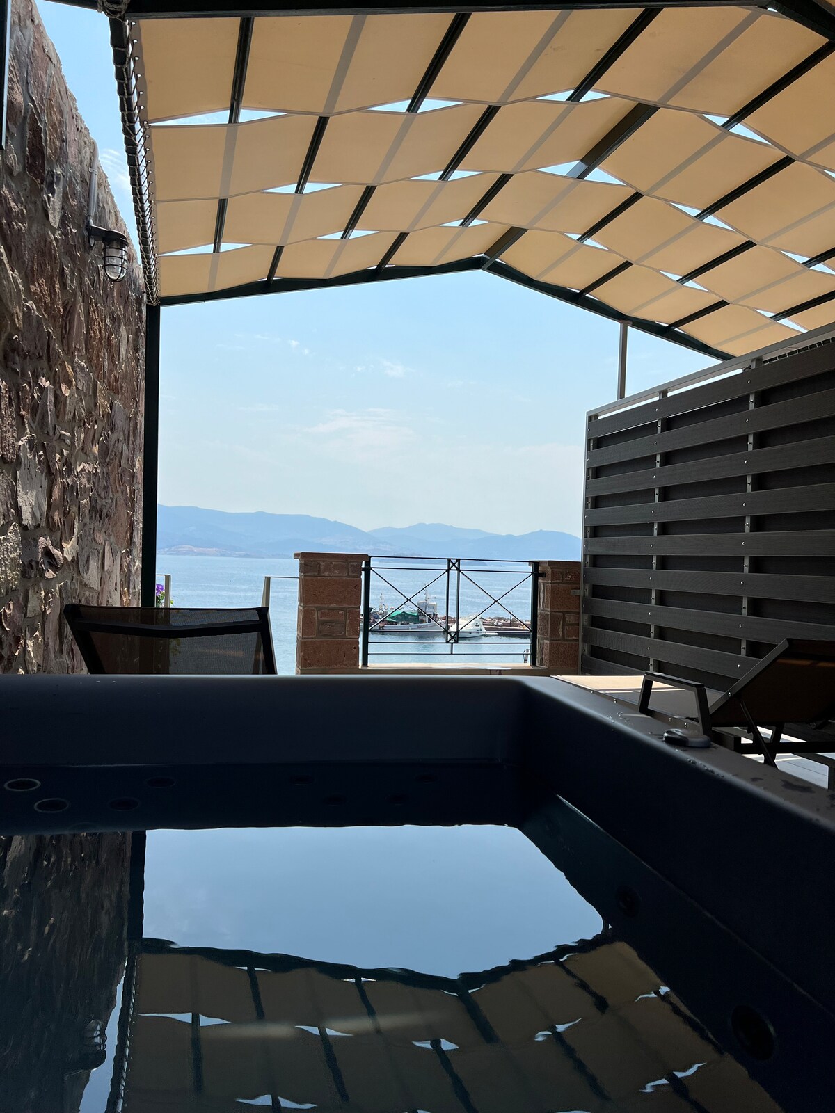A jacuzzi is visible in the foreground, reflecting the serene surroundings. Beyond, a terrace offers views of the Aegean Sea and distant mountains. Lounging chairs are positioned nearby, creating a space for relaxation. A protective awning provides shade over the area.