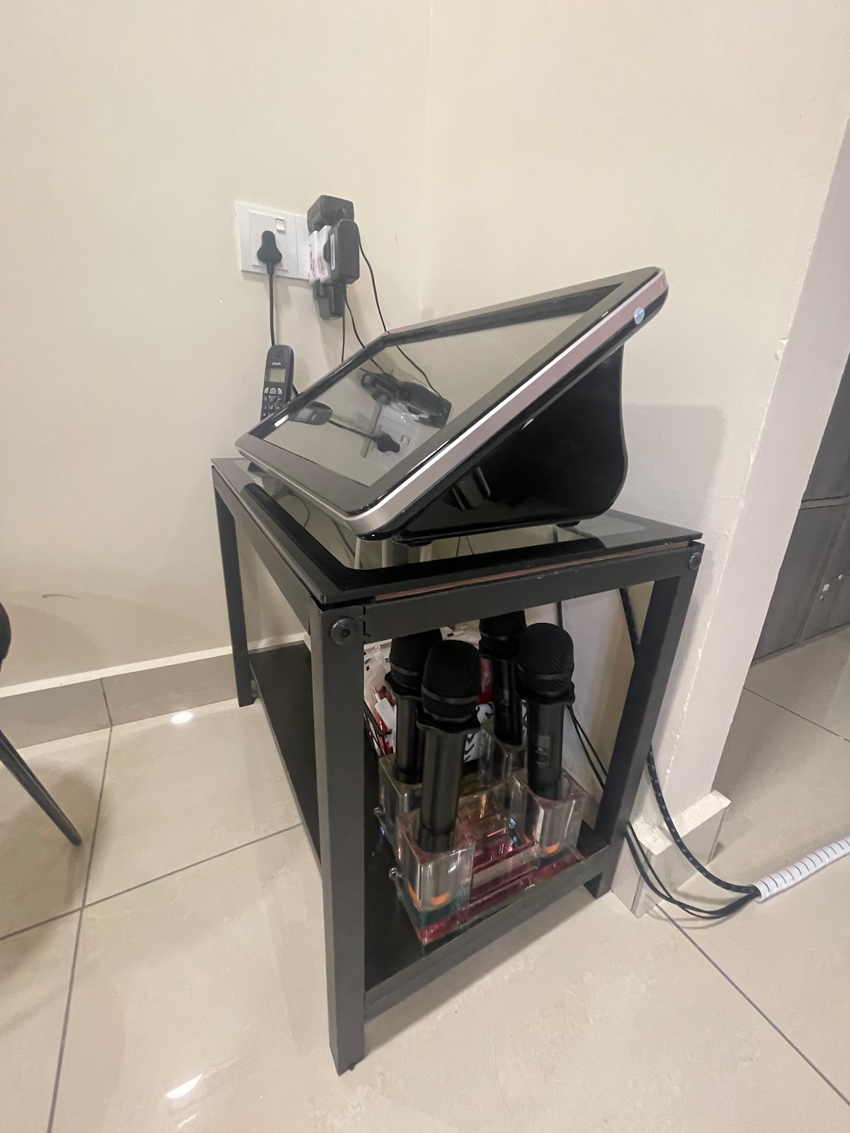 A sleek touchscreen device is positioned on a modern black side table, which also accommodates several microphones stored on a lower shelf. The setup is situated against a neutral wall, with visible electrical outlets and charging devices nearby.