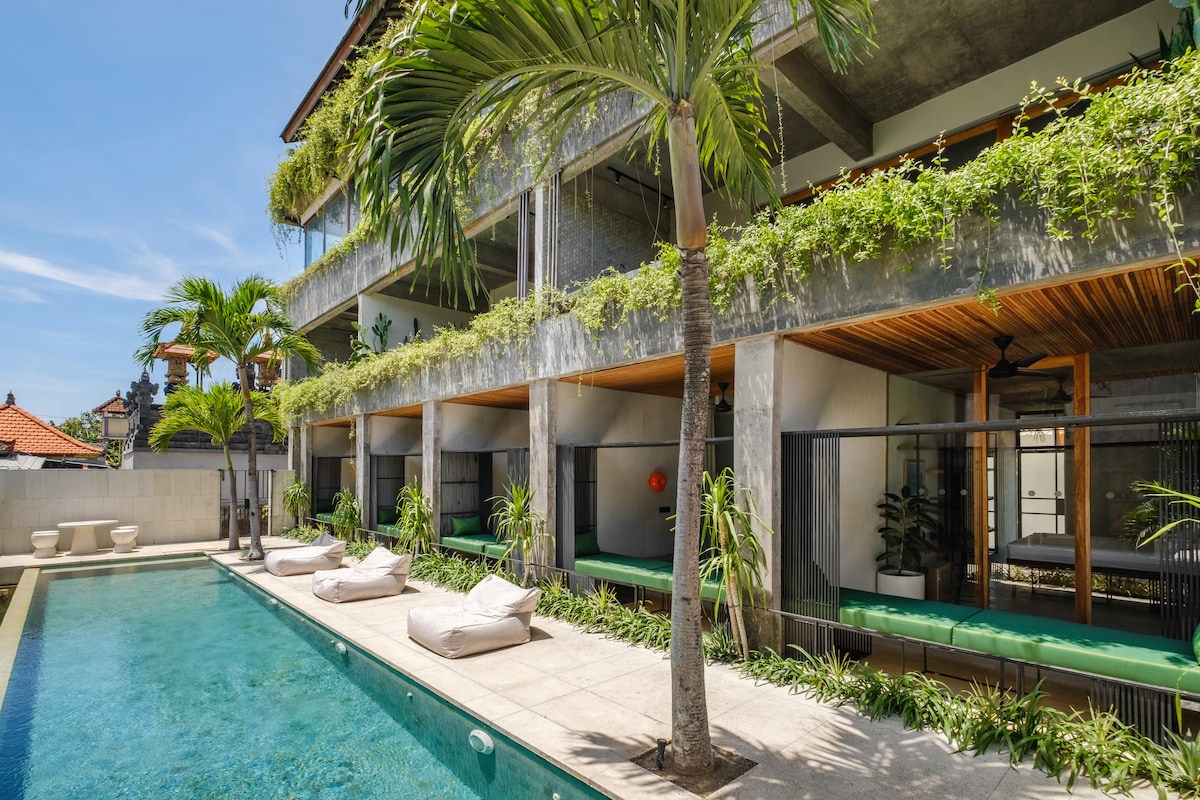 This image displays a modern building featuring a serene swimming pool bordered by lush greenery. Lounge chairs are arranged beside the pool, with large glass doors providing a view into the inviting spaces inside. Palm trees add a tropical touch to the ambiance.