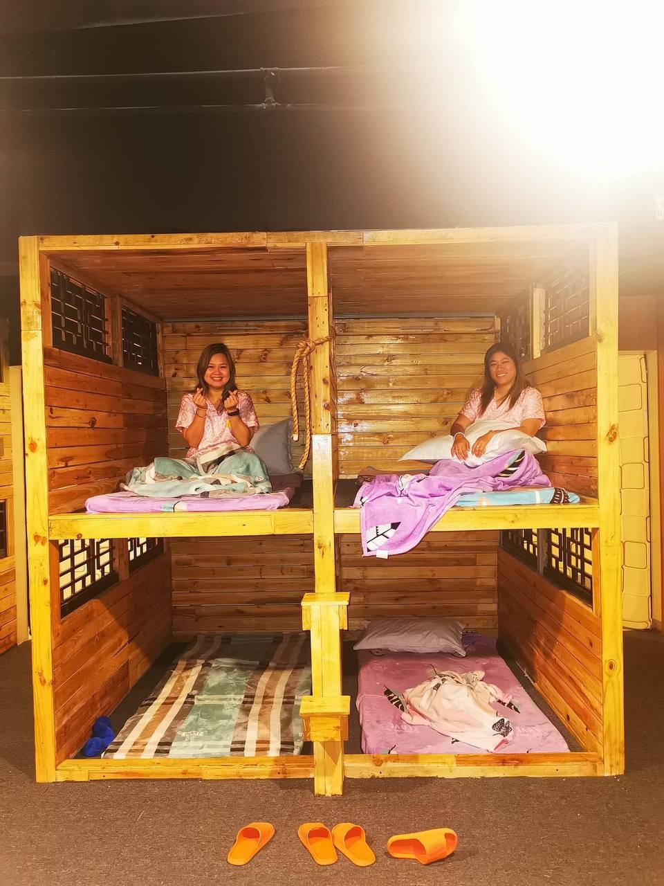 A wooden structure with two separate sleeping areas is seen. Each space features a bunk bed, covered with blankets and pillows. Mats are placed on the floor, and slippers are visible nearby, contributing to a cozy atmosphere.