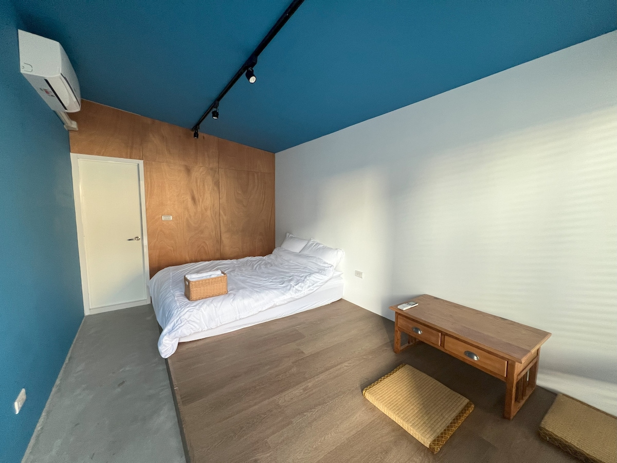 A serene bedroom features a spacious bed with white bedding against a deep blue accent wall. A minimal wooden table and tatami mats complement the natural tones, while a door leads to an adjoining space. Warm light filters through partially covered windows.