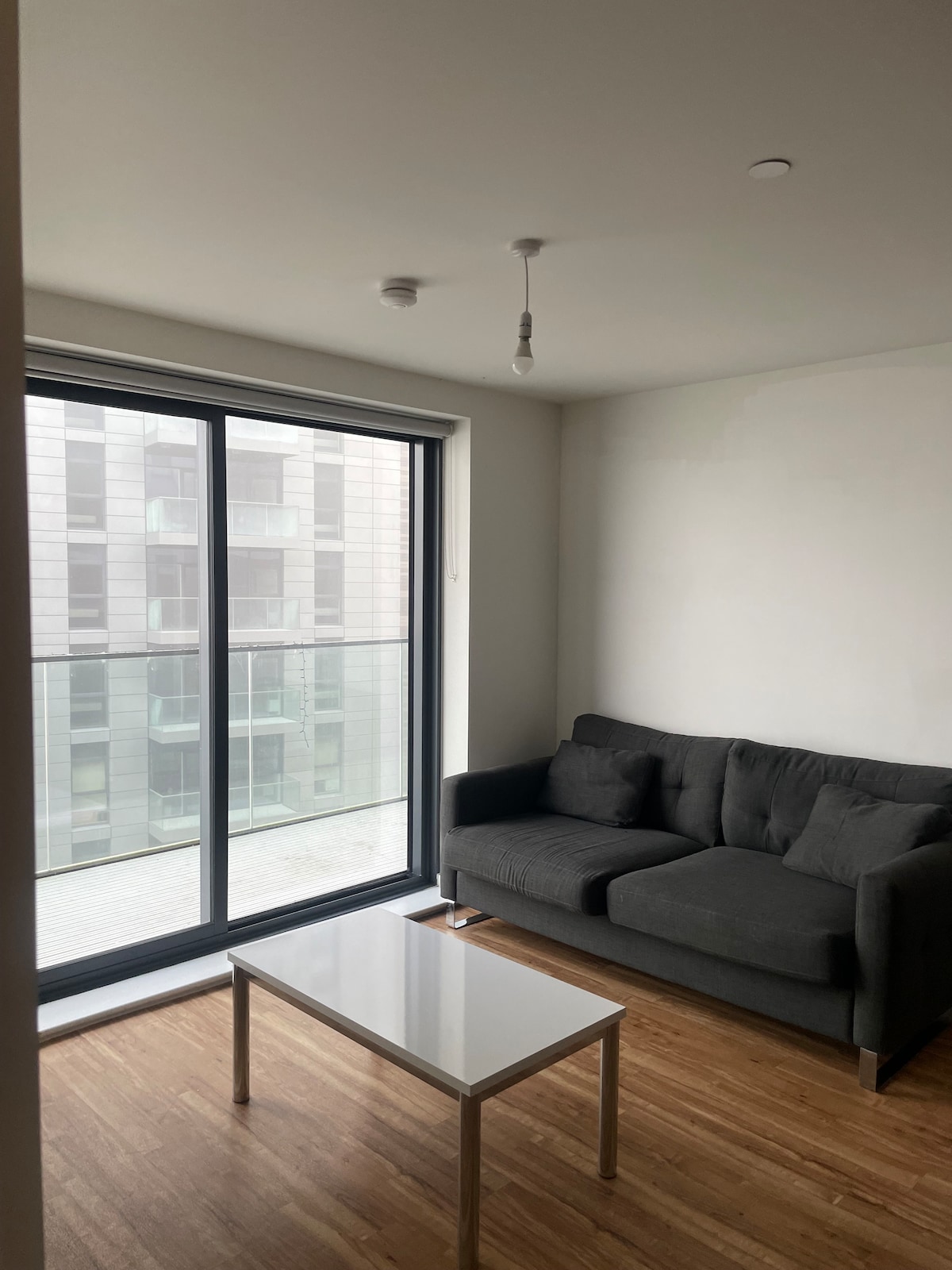 A modern living area features a dark grey couch positioned in front of large glass sliding doors. A small white coffee table is situated centrally on a wooden floor, allowing for ample natural light to illuminate the space.