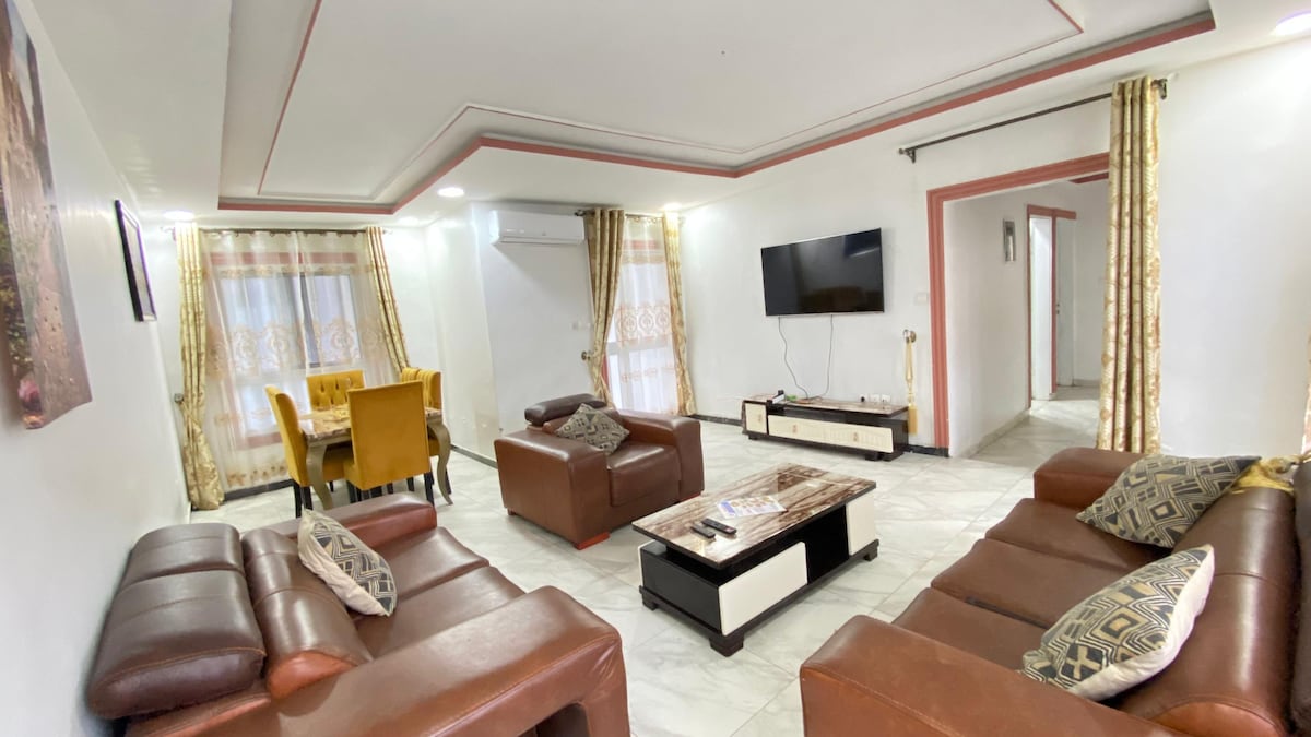 A living area features two brown leather sofas arranged around a coffee table, complemented by patterned throw pillows. A wall-mounted television is visible, with light streaming in through sheer curtains. A dining area with yellow chairs is situated in the background.
