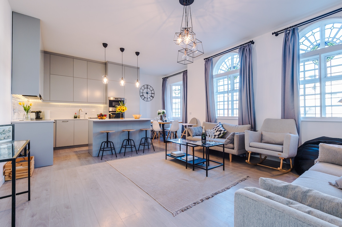 An open living area displays a modern kitchen with pendant lighting and bar stools. A cozy seating arrangement includes light-colored chairs and a sofa, arranged around a low coffee table. Large windows allow natural light to fill the space, enhancing the inviting atmosphere.