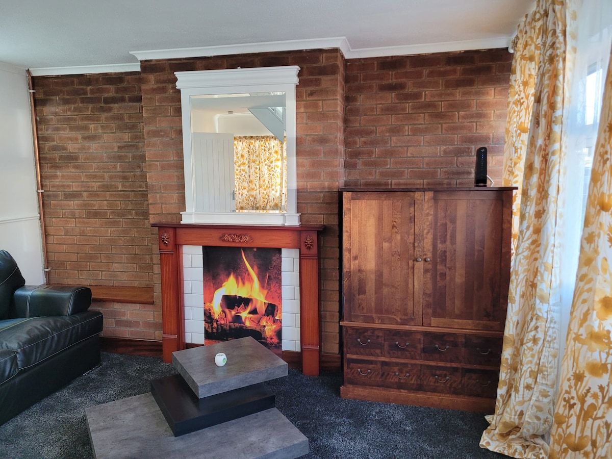 A cozy living area features a traditional fireplace with a visible flame. A large wooden cabinet stands beside it, and a low coffee table rests on a plush carpet. Sunlight filters through vibrant yellow curtains, adding warmth to the space.
