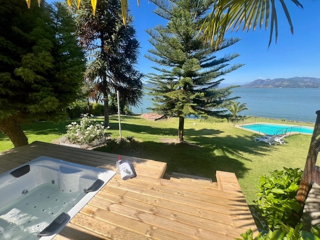 A wooden deck surrounds a modern jacuzzi, providing views of the expansive garden and lake. Lush green grass is complemented by palm and conifer trees. A swimming pool is visible in the background, along with lounge chairs for relaxation.