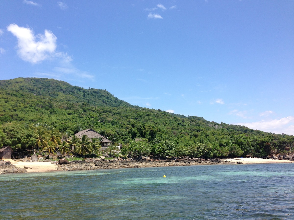 A lush landscape is depicted, featuring a mix of greenery and palm trees along the shoreline. A beach sits adjacent to the tranquil water, while a modest structure is visible amidst the natural surroundings, framed by the mountainous backdrop under a clear blue sky.
