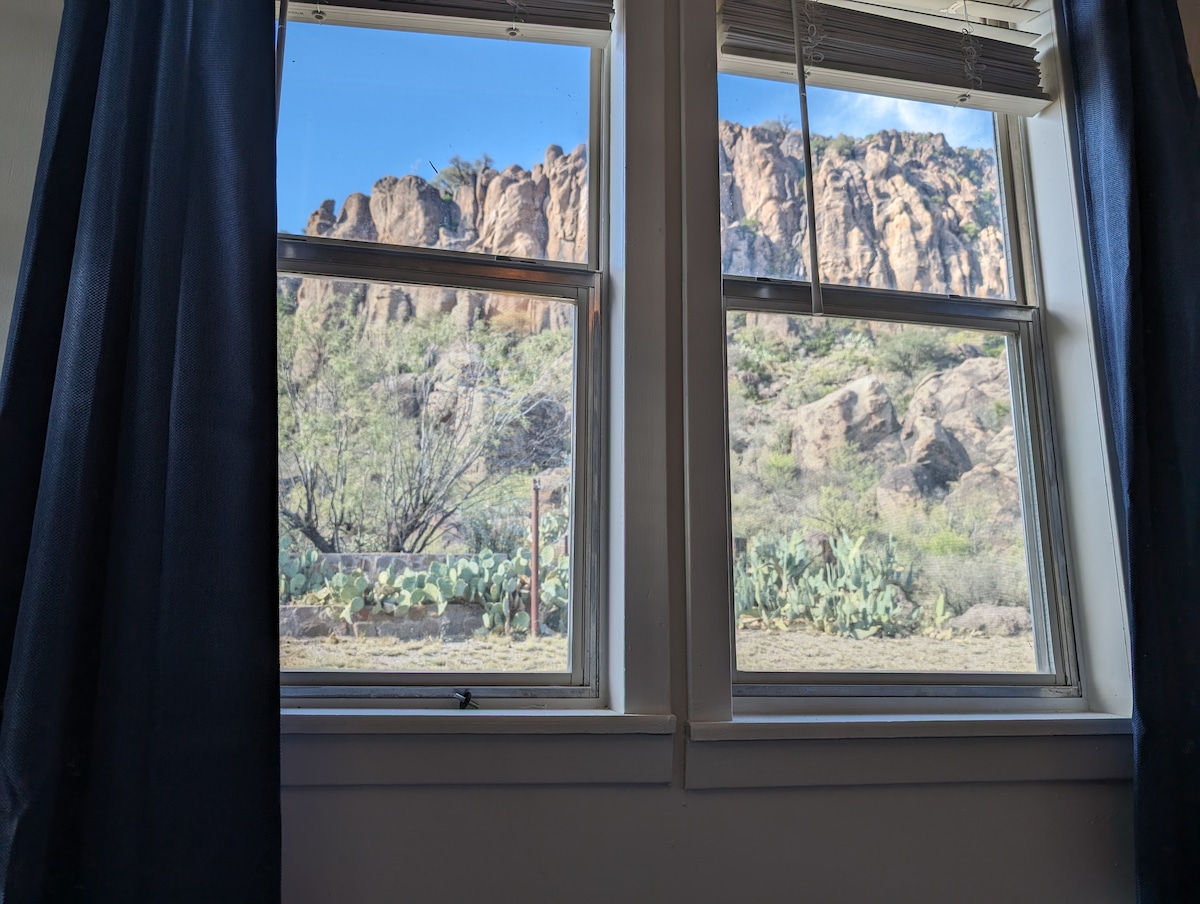 A view is captured through two framed windows, showcasing a rugged rock face and surrounding greenery. Sunlight illuminates the landscape, highlighting the textures of the rocks and the vibrant plants below. Dark curtains partially obscure the scene, creating a cozy and sheltered atmosphere.