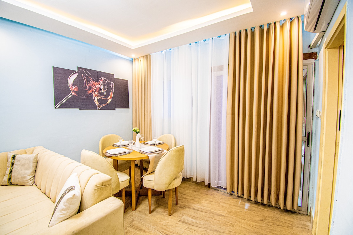 A dining area is visible, featuring a round table set for four. Light wood dining chairs surround the table. Soft curtains cover the window, allowing filtered light to enter. A piece of wall art hangs above the table, adding a touch of elegance to the space.