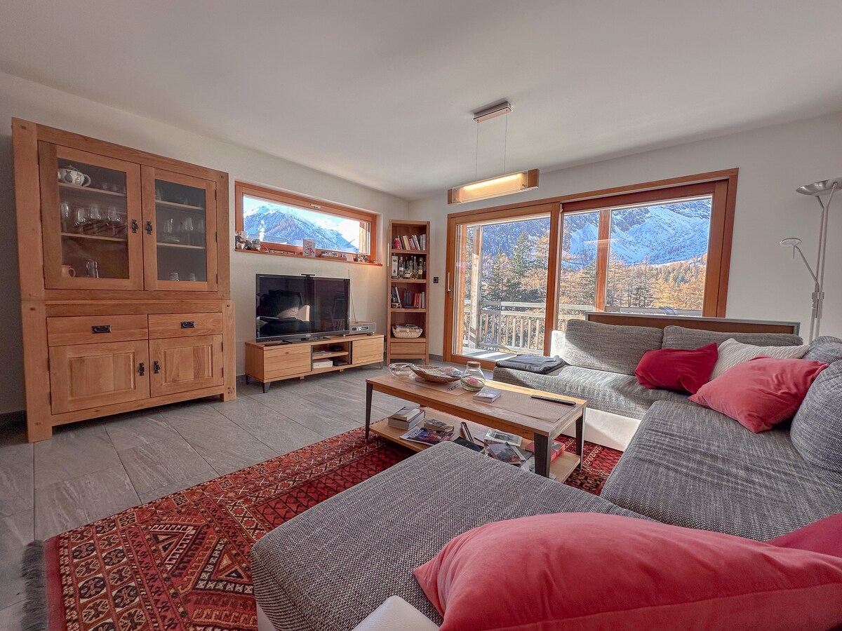 A spacious living area features large windows with mountain views. Comfortable seating is arranged around a low coffee table, accented by a vibrant rug. A television is located on a wooden stand, and shelves display books and decorative items, creating an inviting environment.