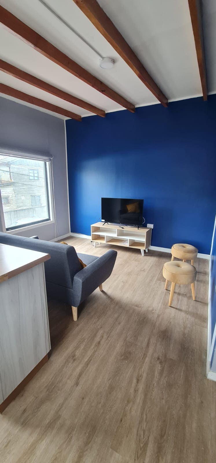 A living area features a blue accent wall and wooden beams. A television is positioned on a light-colored media unit. A cozy chair is placed nearby, alongside a small stool. Natural light enters through a window, enhancing the simplicity of the space.