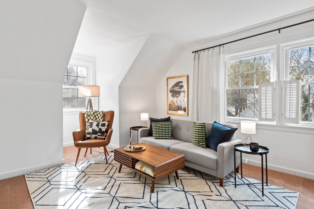 A bright living room features a modern gray sofa adorned with decorative pillows and a coffee table at its center. A wooden armchair with a patterned cushion is positioned beside a tall lamp. Large windows invite natural light, complemented by sheer curtains and a framed artwork on the wall.