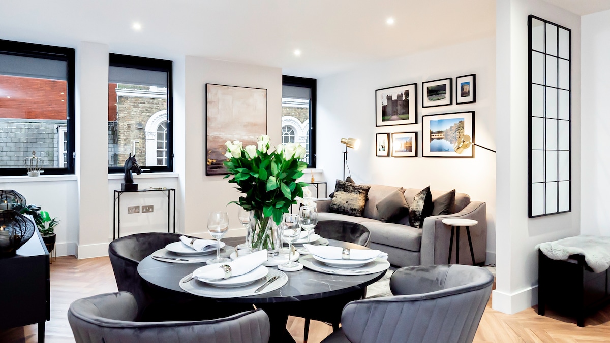A sophisticated living area features a round dining table set for four, complemented by plush seating arrangements. Natural light floods the space through three large windows. The walls are adorned with curated art pieces, enhancing the elegant ambience of the room.