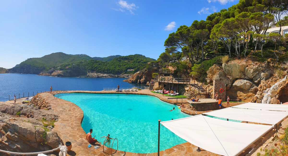 A large salt-water pool is nestled among rocky cliffs, surrounded by lush green trees. The serene water reflects the blue sky, while several sun loungers are positioned along the pool's edge. Guests can be seen enjoying the relaxing atmosphere.