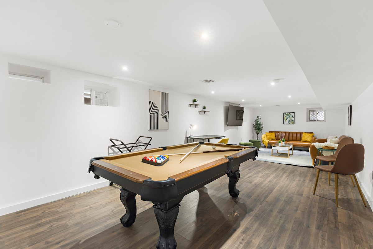 A spacious game area is featured, highlighted by a classic pool table at the center. Surrounding seating includes a comfortable sofa and stylish chairs. Bright overhead lights illuminate the space, and large windows allow natural light to enhance the inviting atmosphere.