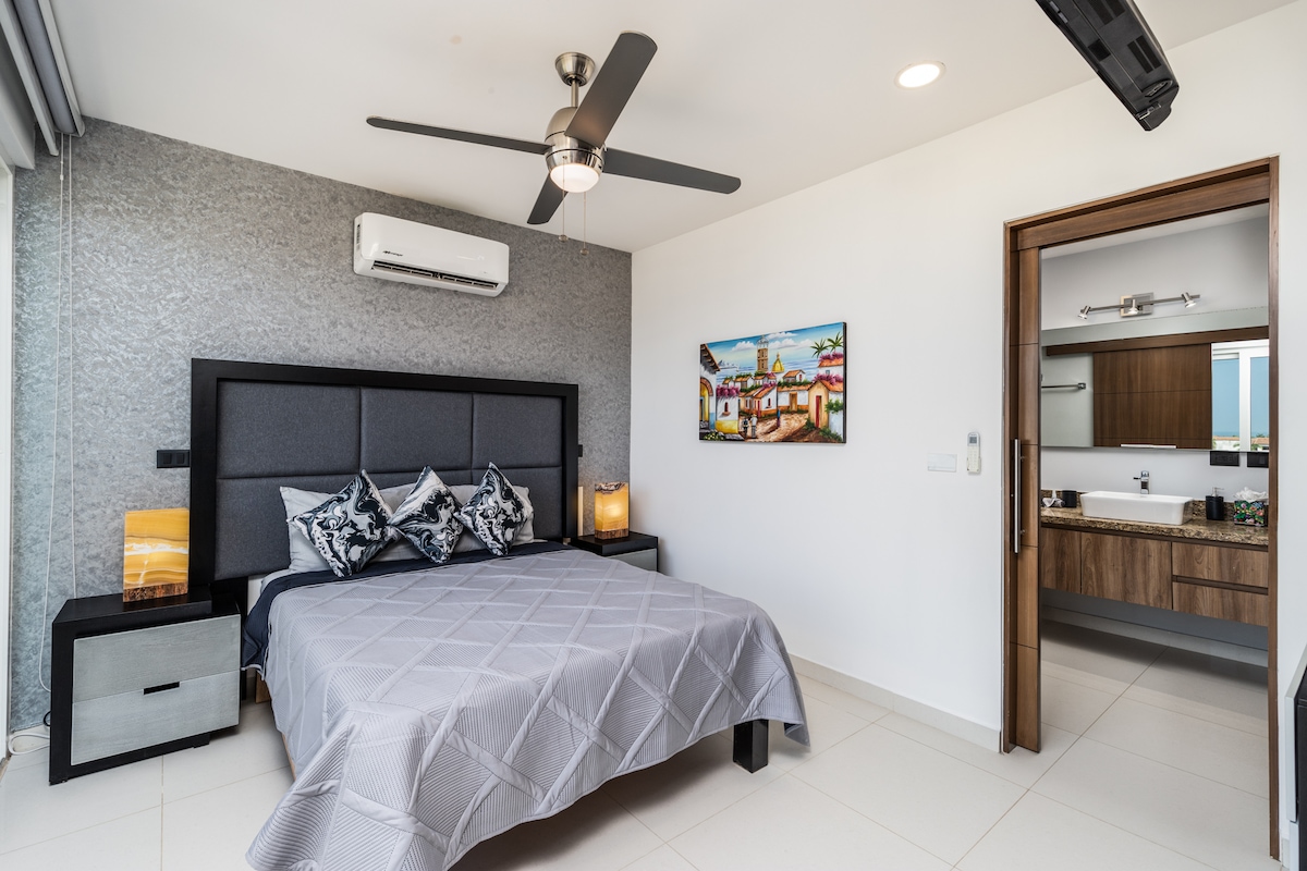 A modern bedroom is highlighted by a spacious bed with a textured gray blanket. Two decorative pillows adorn the bed, complemented by matching bedside lamps. A wall-mounted fan provides airflow, and an artwork depicting a colorful scene adds visual interest. Natural light enters through large windows.