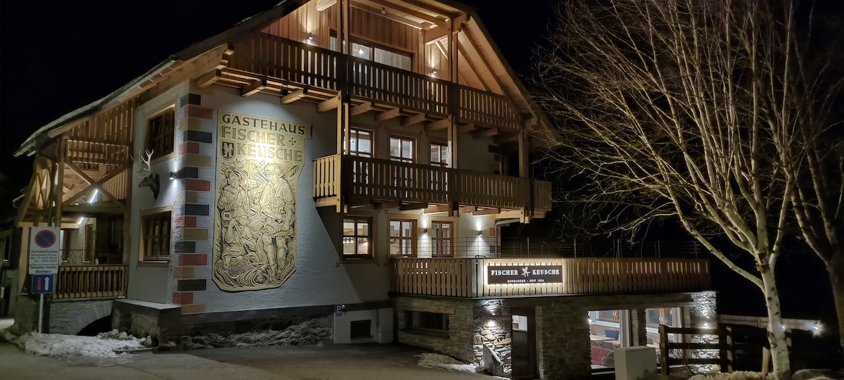 The exterior of the building is illuminated at night, showcasing wooden balconies and detailed carvings on the wall. Soft lighting highlights the architectural features and provides a welcoming ambiance. The entrance area and surrounding landscape are partially visible.