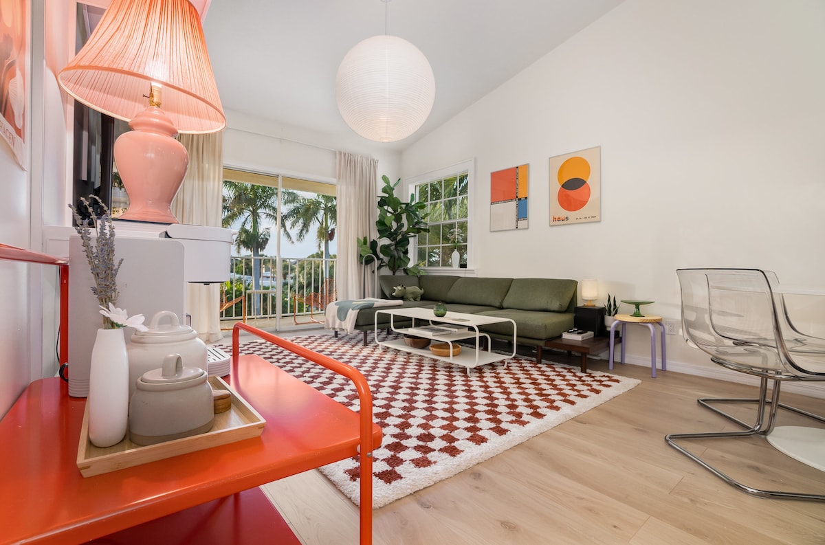 A spacious living area features large windows allowing natural light to fill the room. Stylish furnishings include a green sofa, a round coffee table, and modern chairs. A decorative rug adds texture, while tropical plants and artwork contribute to a contemporary aesthetic.