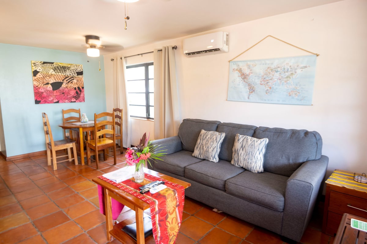 A cozy living area is featured with a gray sofa adorned with decorative pillows. A small wooden table and chairs are visible in the background. Natural light enters through a window covered by light curtains, while a colorful table runner adds a touch of warmth.