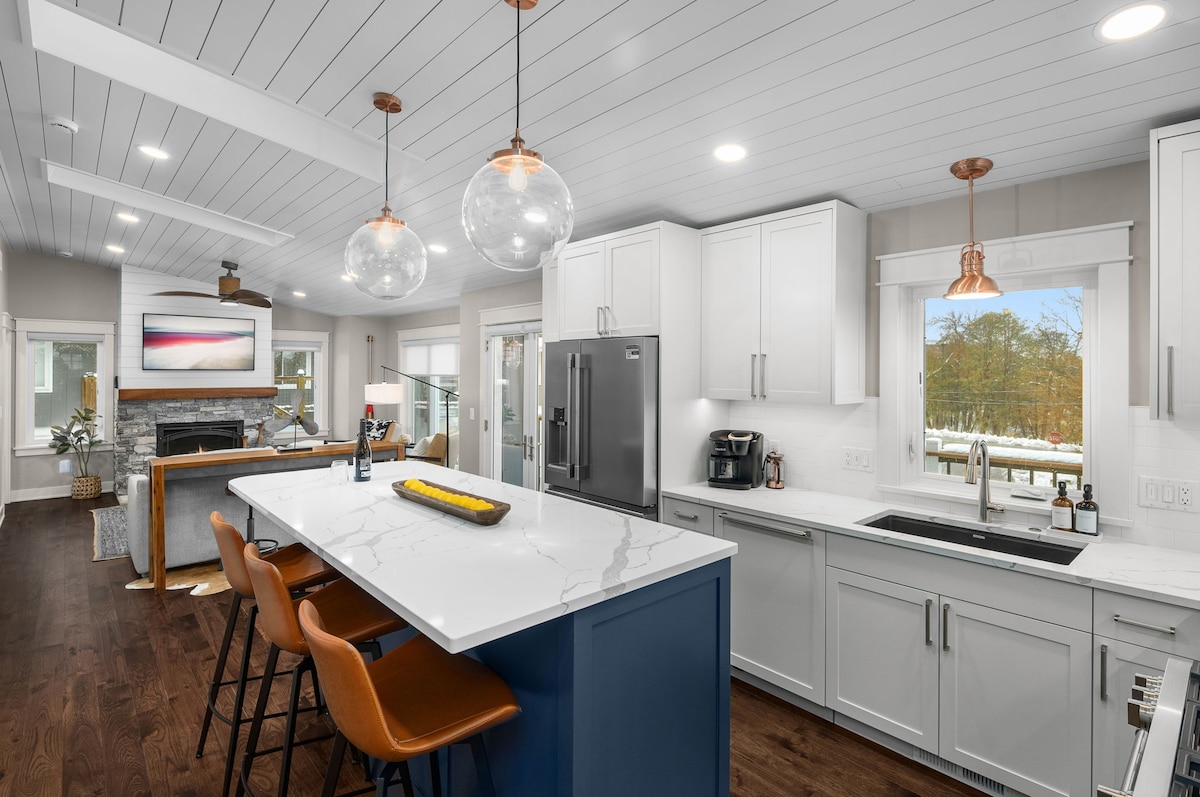 The kitchen is designed with modern finishes, featuring a central island with seating for four. Stainless steel appliances complement the white cabinetry, while large windows provide natural light and views of the outdoor area. The open layout connects seamlessly to the living space, enhancing flow.