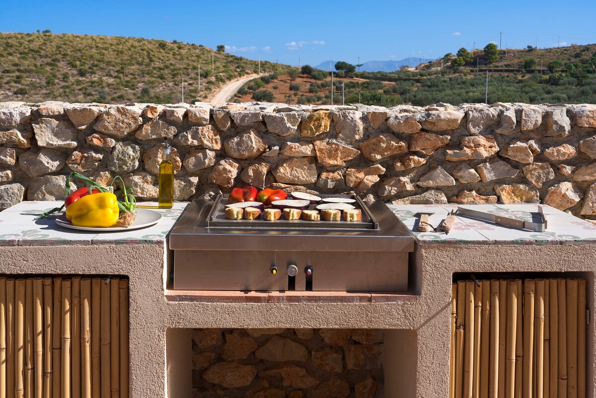 An outdoor cooking area constructed from stone features a gas barbecue, with vegetables and olive oil arranged on the countertop. The surrounding landscape includes rolling hills and a clear blue sky, providing a scenic backdrop for outdoor dining.
