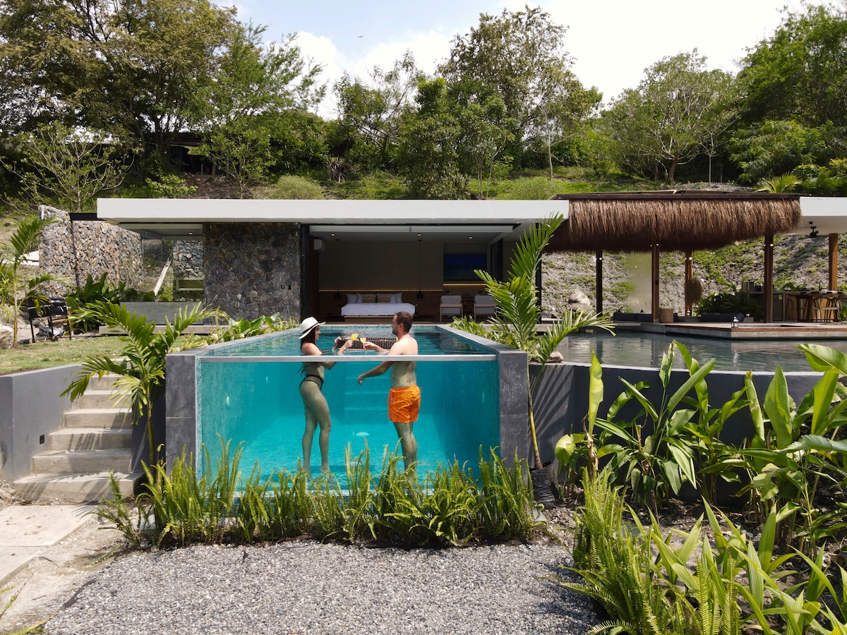 A modern outdoor pool area is surrounded by tropical greenery, featuring two guests enjoying the inviting water. A covered seating area with natural materials is visible in the background, nestled against a stone wall and complemented by additional lounge spaces.