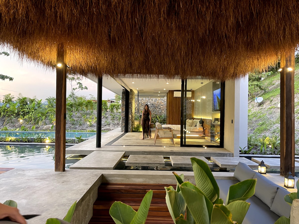 A spacious outdoor area features a straw-covered overhang, framing a view of the modern indoor living space through large glass doors. The patio is bordered by lush greenery and a serene pool reflects the surroundings, creating a tranquil atmosphere.