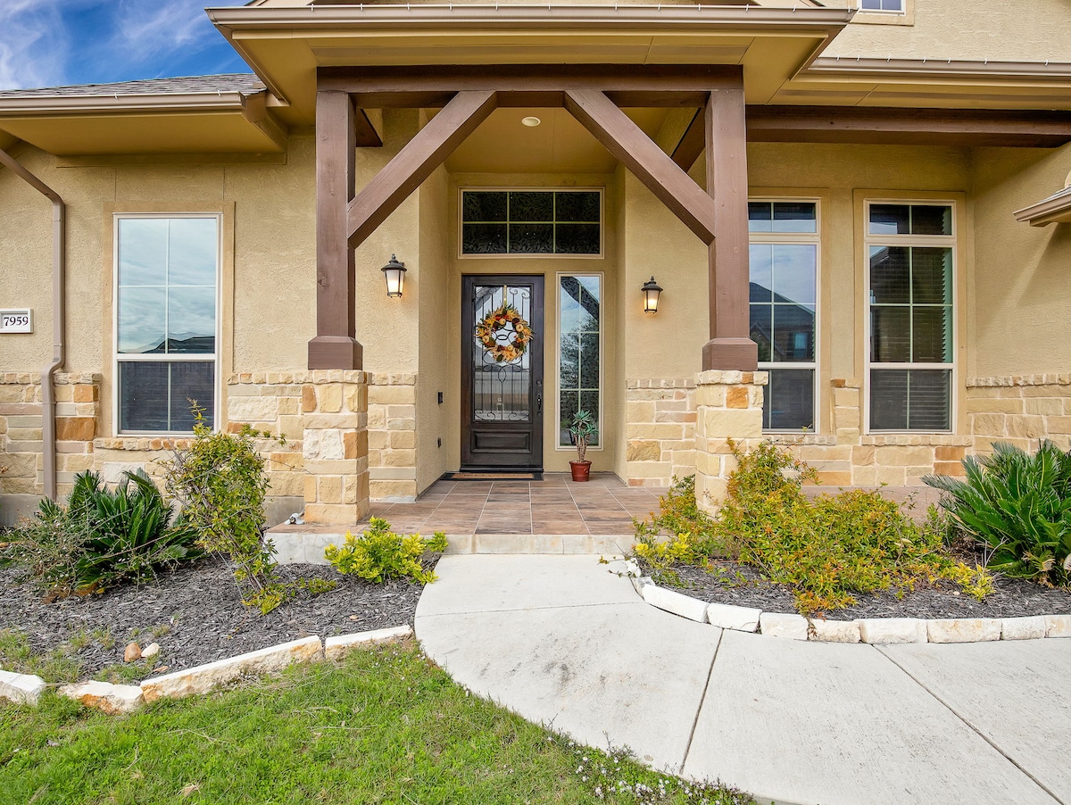 The entrance showcases a welcoming front porch with large stone columns and a stylish wooden door. Flanking the door are elegant wall sconces, and a seasonal wreath adds a touch of charm. Well-maintained landscaping features shrubs and flowering plants along the walkway.