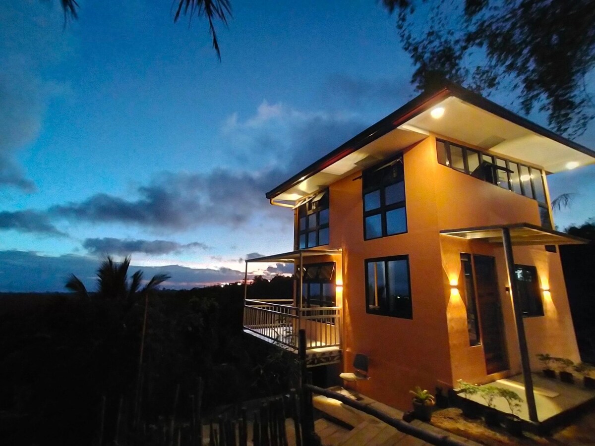 A modern two-story house is seen against a twilight sky, featuring large windows that allow views of the surrounding landscape. Soft lighting is provided by exterior fixtures, highlighting the building's exterior. A wooden pathway leads to the entrance, accompanied by greenery.