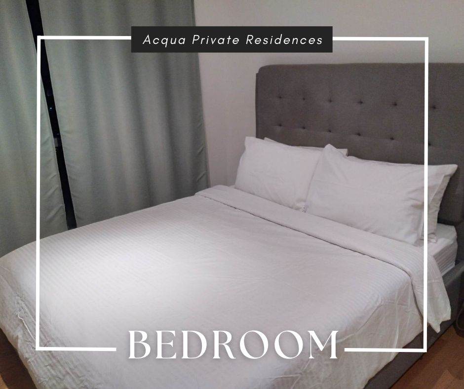 A bedroom features a spacious bed with white bedding and a gray upholstered headboard. Soft gray curtains frame a window, allowing natural light to illuminate the room's neutral tones. The wooden floor adds warmth to the overall atmosphere.