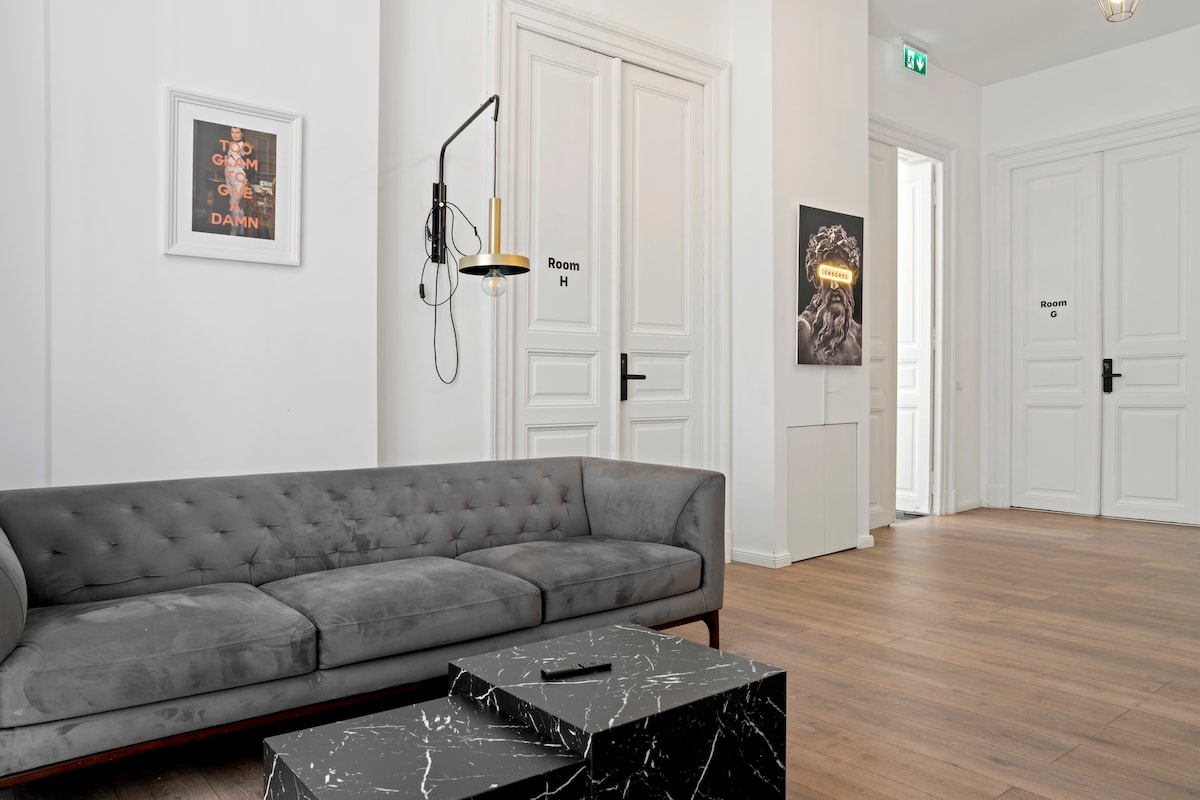 A modern common area features a plush gray sofa and sleek black marble coffee table. Light streams in through large doors, highlighting the minimalist decor. Room signs are visible on the doors, contributing to a sense of accessibility and organization in the space.