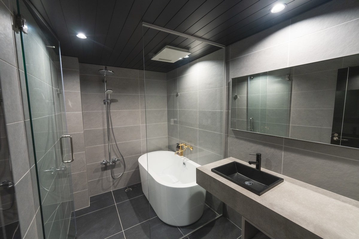 A modern bathroom features a freestanding white bathtub and a walk-in shower with glass doors. A dark stone sink rests on a counter of gray material. Large mirrors are placed above the sink, and overhead lights provide ample illumination.